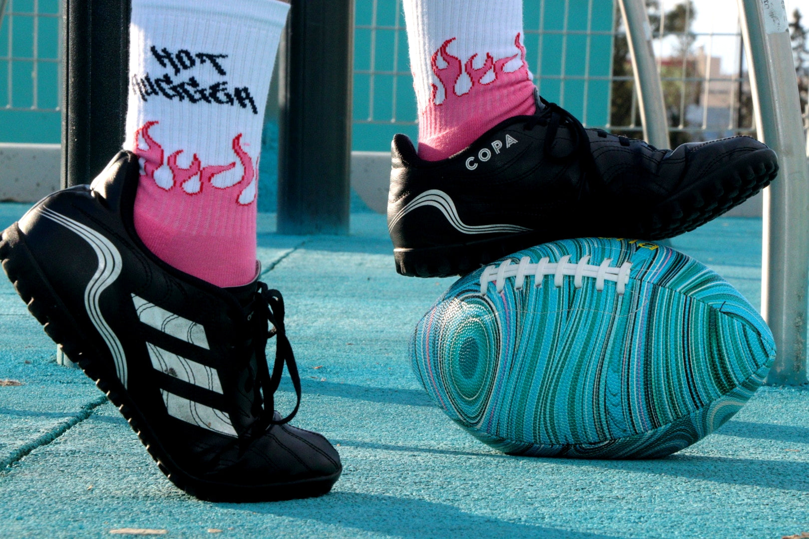 Person wearing black Adidas shoes with pink socks stepping on a blue and green striped ball.