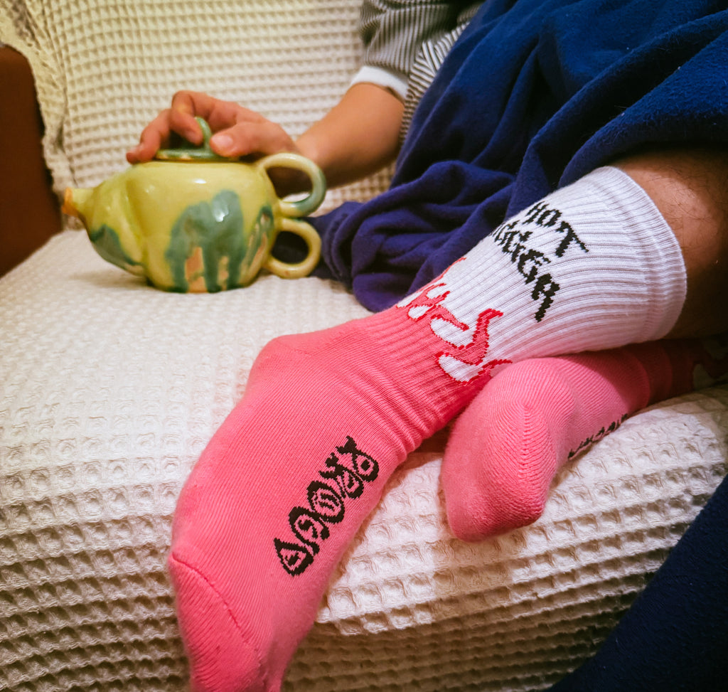 Person wearing pink socks with text, sitting on a textured chair holding a green teapot.