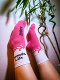 Pink socks with black text and white accents worn by a person, with a plant in the background.