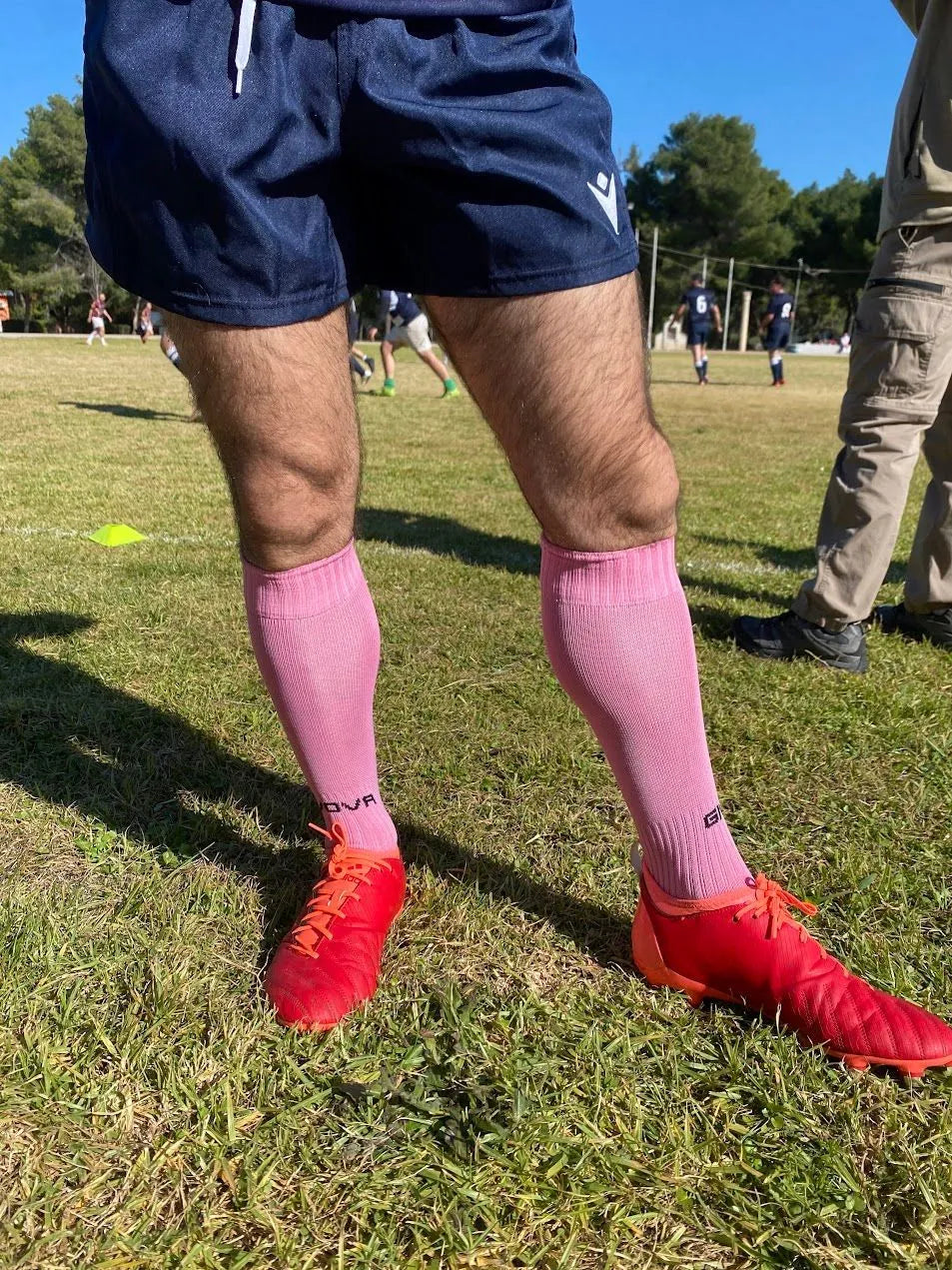 Person wearing pink socks and red cleats on a grassy field with other people in the background.
