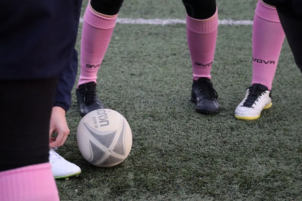 Person wearing pink socks and black shoes with a rugby ball on a grass field