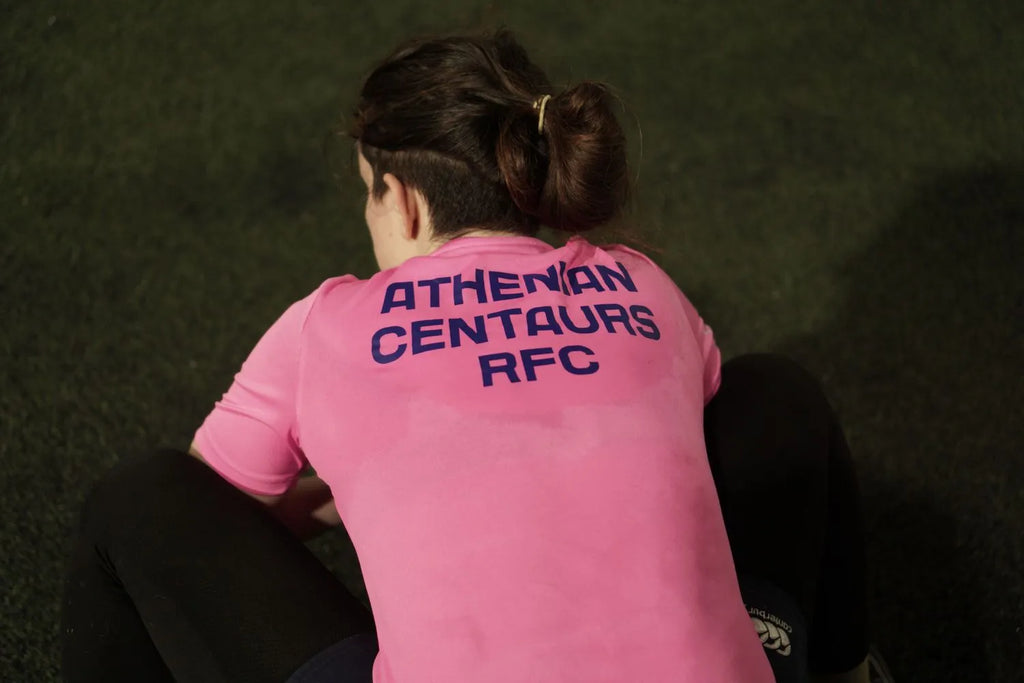 Person wearing a pink shirt with 'Athenian Centaurs RFC' text, sitting on grass.