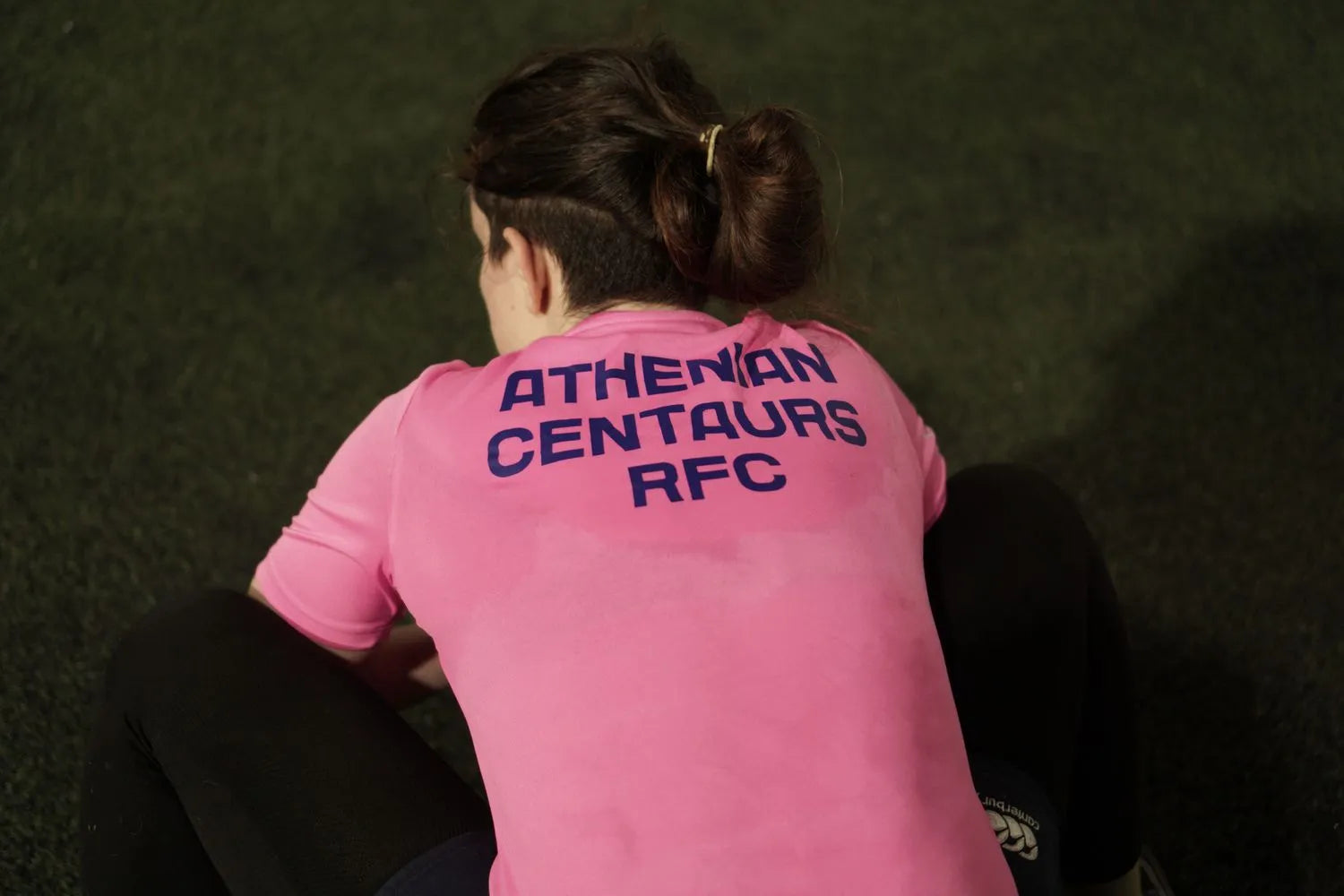Person wearing a pink shirt with 'Athenian Centaurs RFC' text, sitting on grass.